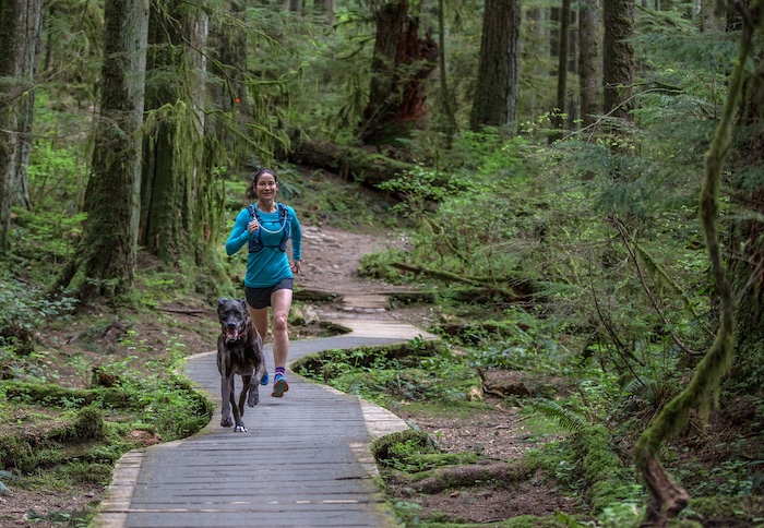 A woman runs along a planked pathway through a redwood forest with her dog.