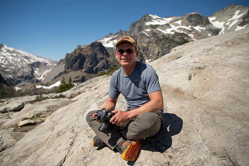 Jason Hummel smiling and sitting on a rock with his camera