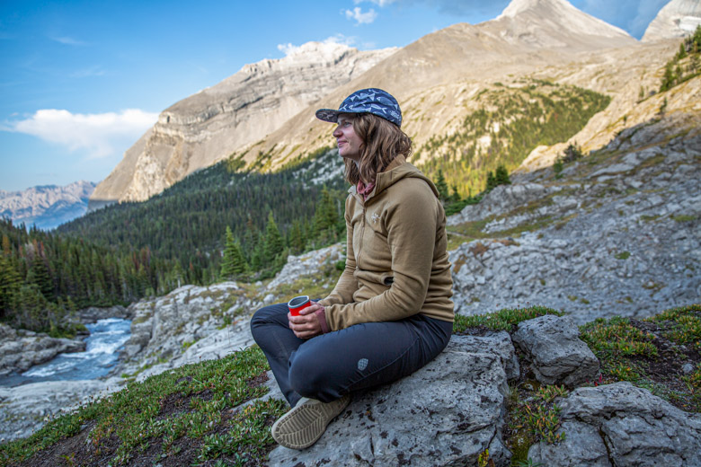 A woman sitting on a rock in a mountainous landcape wearing a brown fleece jacket