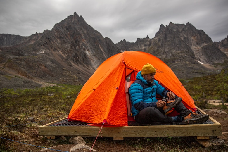 A man puts on shoes in his tent while camping in front of scenic mountains