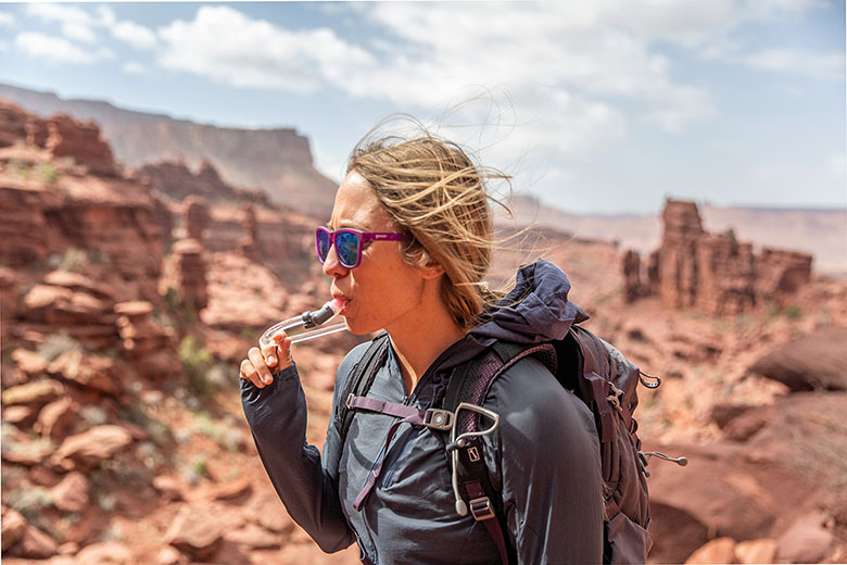 A woman drinks from a hydration backpack while hiking in the desert.