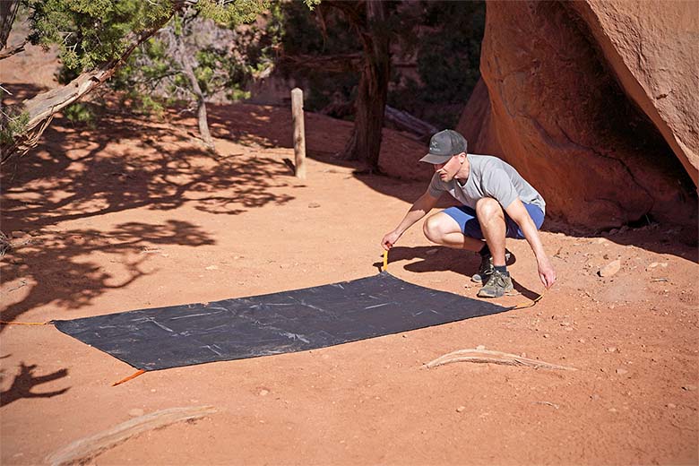 A man lays down a tent footprint on dusty desert ground.