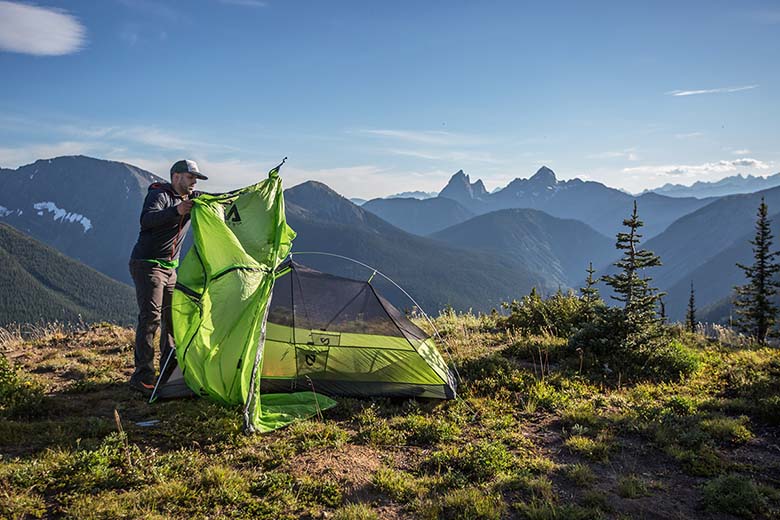 A man puts the rainfly on a green tent with mountains on the horizon