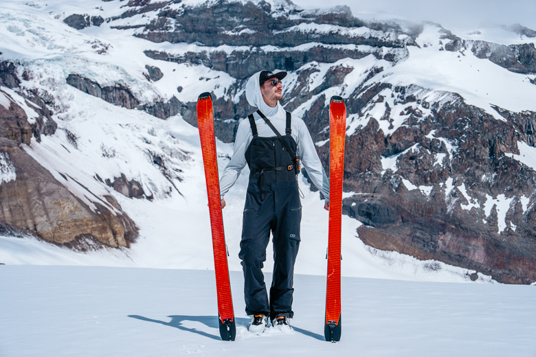 A man wearing ski bibs is standing with skis in front of glacier