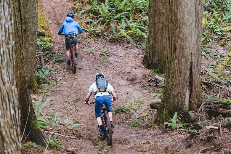 Two mountain bikers bike up a hill, one with a backpack and one with a waist pack