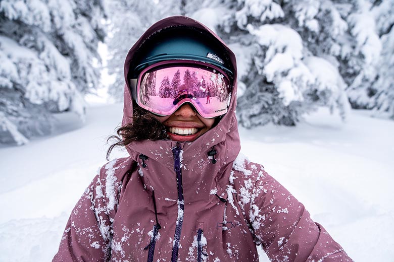 A smiling woman wears Oakley goggles while surrounded by snow and trees