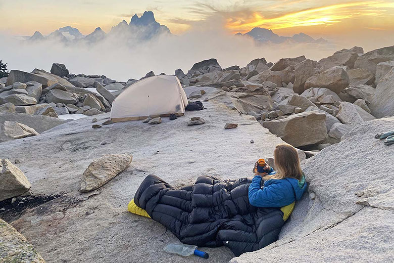 A woman sits against a rock in a sleeping bag, overlooking a foggy mountain landscape