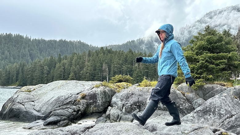 A woman wearing rain boots and a rain jacket walks along rocks on a cloudy day