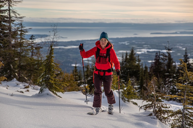 A woman skins up a snowy hill on skis wearing a red synthetic jacket