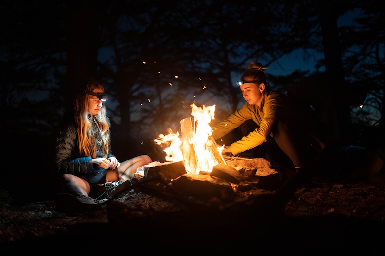 Two women sit in the dark around a large campfire