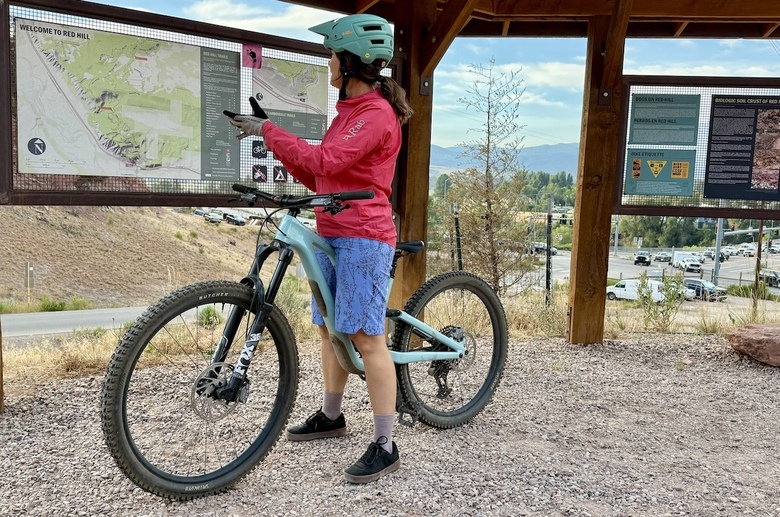 A woman stands at the trailhead for mountain biking in the Rab Cinder Phantom rain jacket