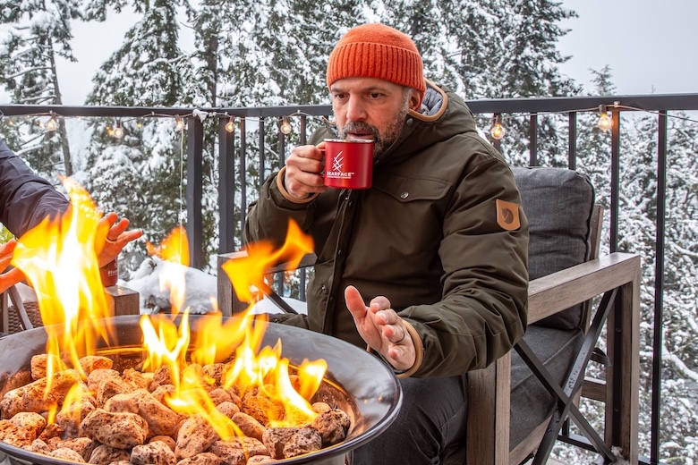 A man sits next to a fire pit on a snowy day, warming his hands