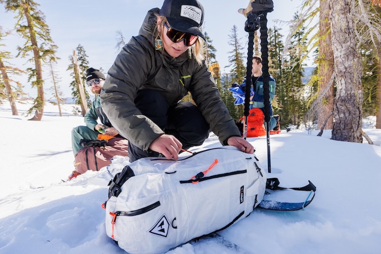 A skier kneeling down in the snow unzips a backpack