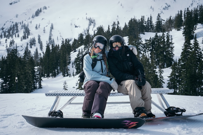Two snowboarders sit smiling on a bench