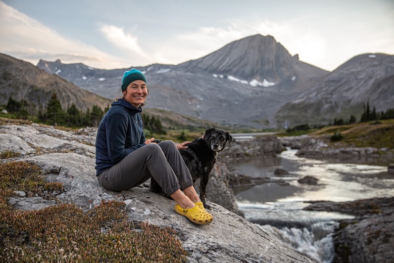 A woman sits on a rock in front of a mountain range with her dog