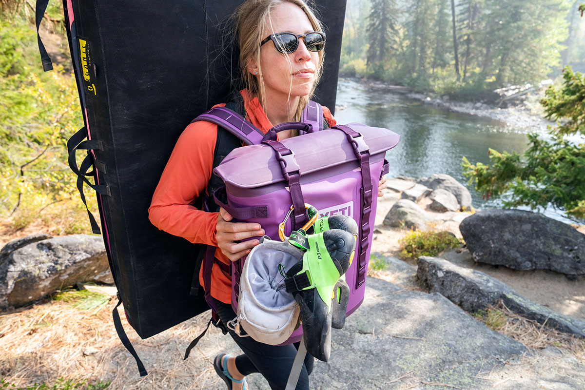 A climber carries a crashpad on her back, and a backpack cooler on her front, to pack out all her gear for the day.