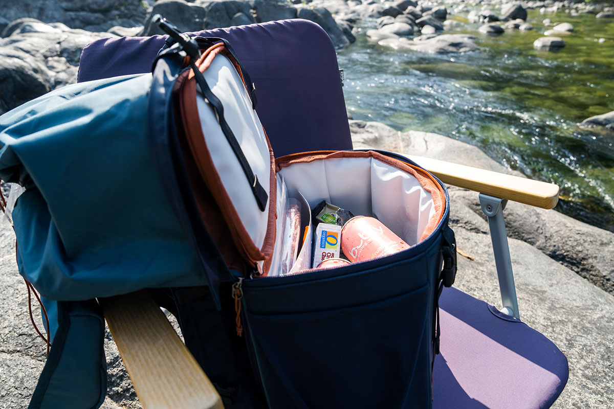 A backpack cooler is perched on a beach chair next to a river.