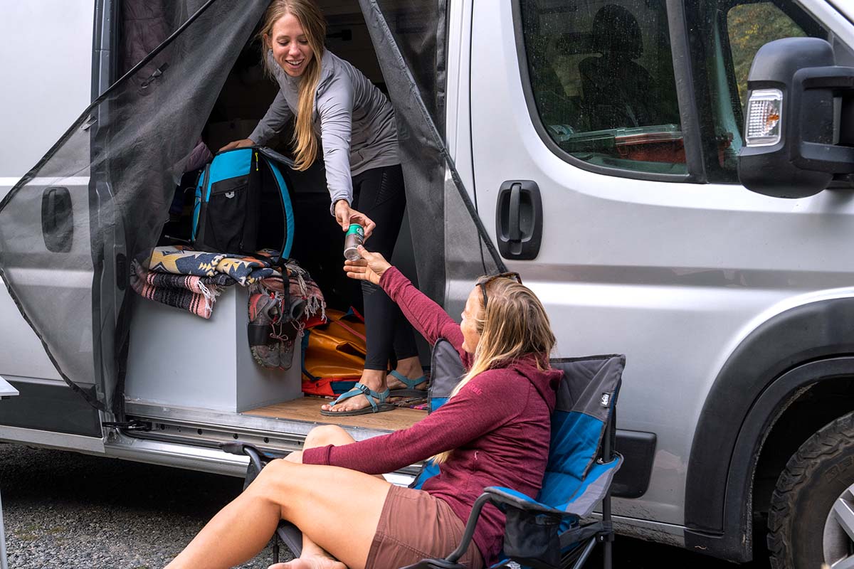A woman hands a drink from her cooler to a friend sitting nearby in a chair.