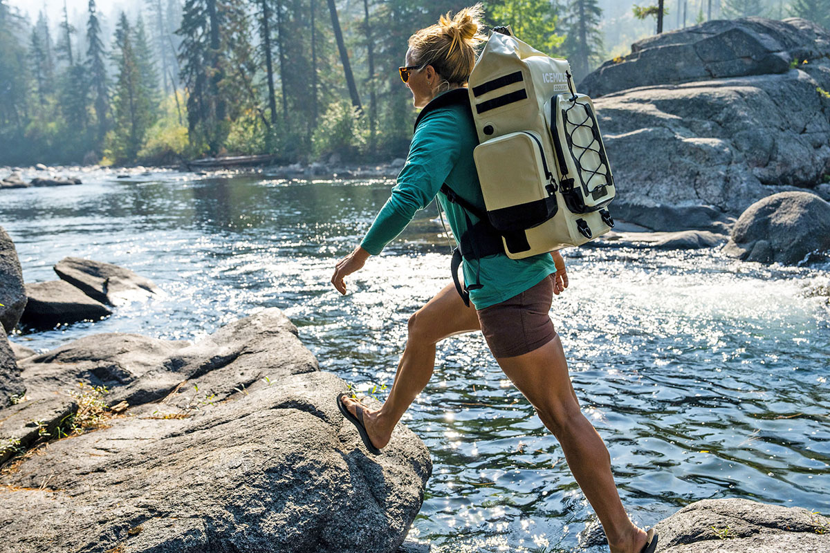 A hiker hops on a rock while crossing a lake with her Icemule backpack cooler.