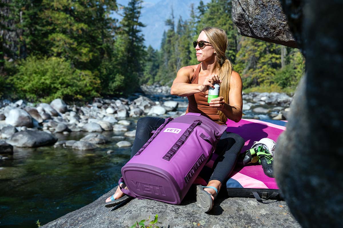 A woman sits on a rock next to a swimming hole and pulls a drink from her backpack cooler.