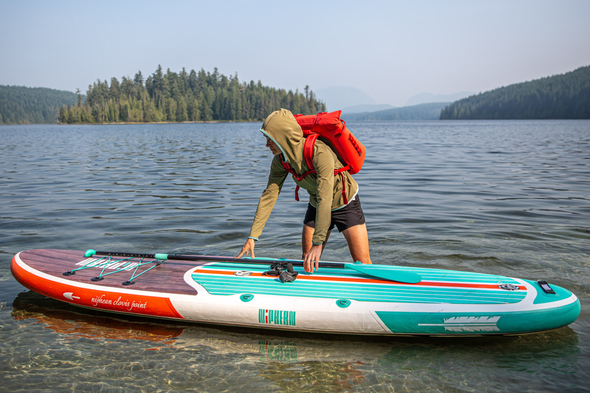A woman standing in the water with a paddle board and backpack cooler