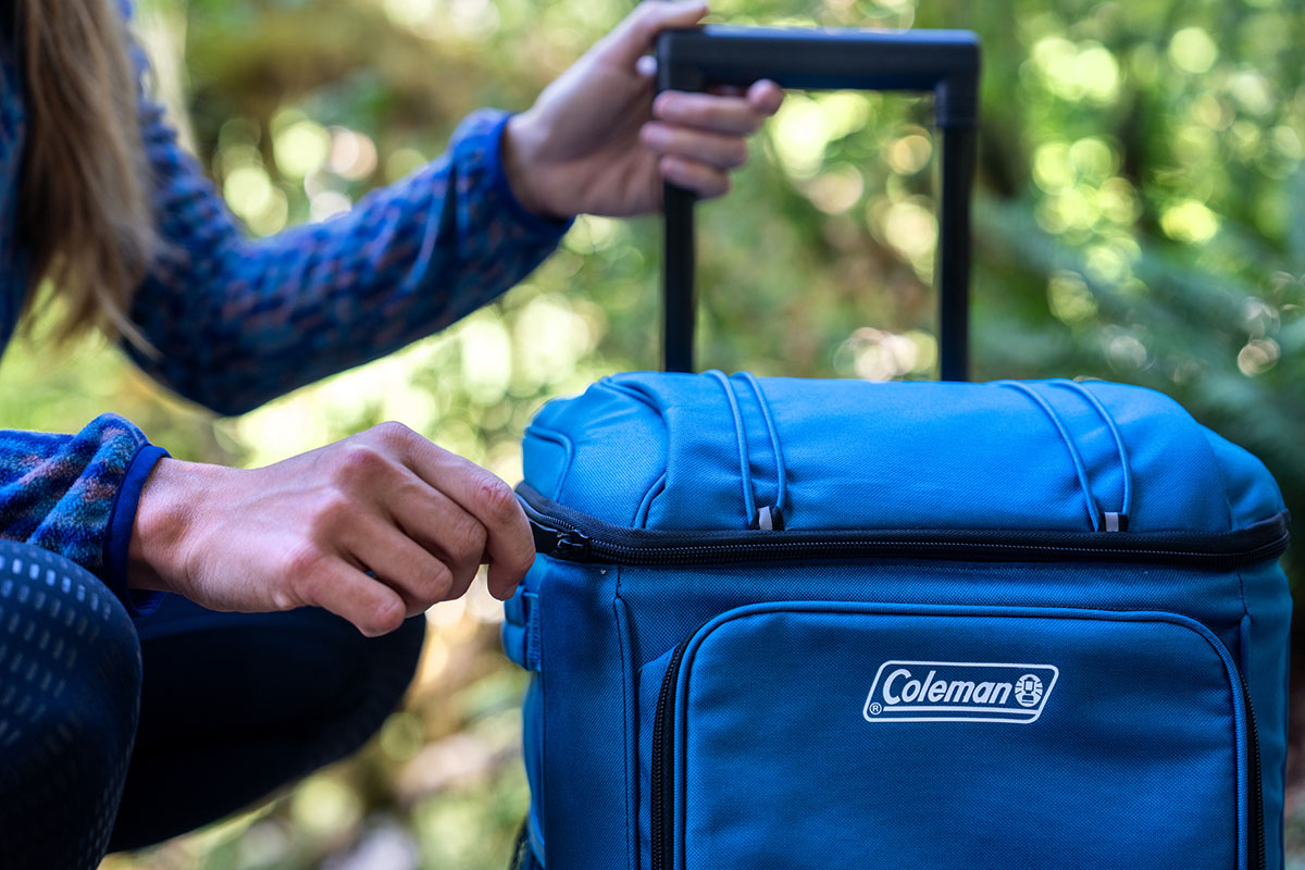 A camper unzips the lid on a wheeled Coleman cooler.