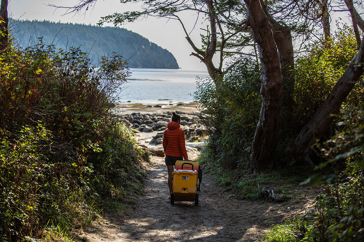 A woman rolls a cooler down a dirt path to a rocky lake shoreline.