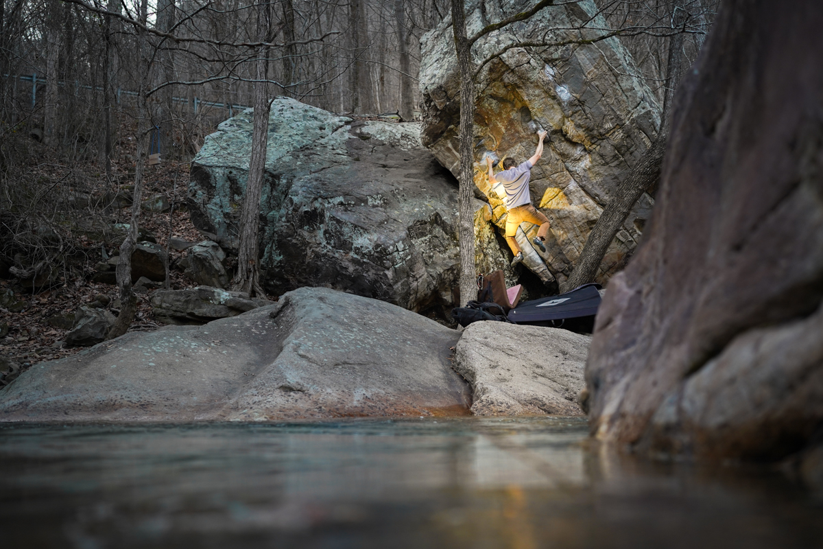 A man climbing a boulder near a river at dusk with bouldering crash pads