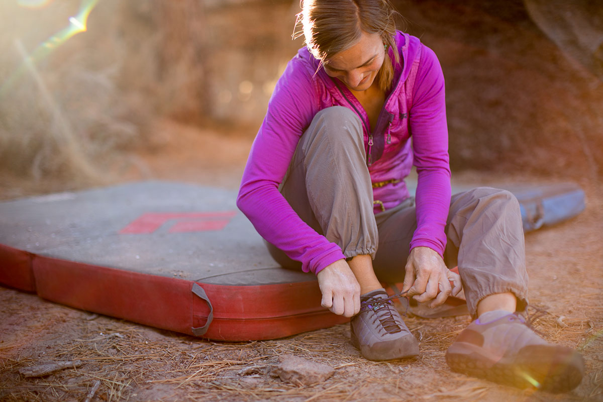 A woman sit on the edge of a bouldering crash pad while she ties her climbing shoes