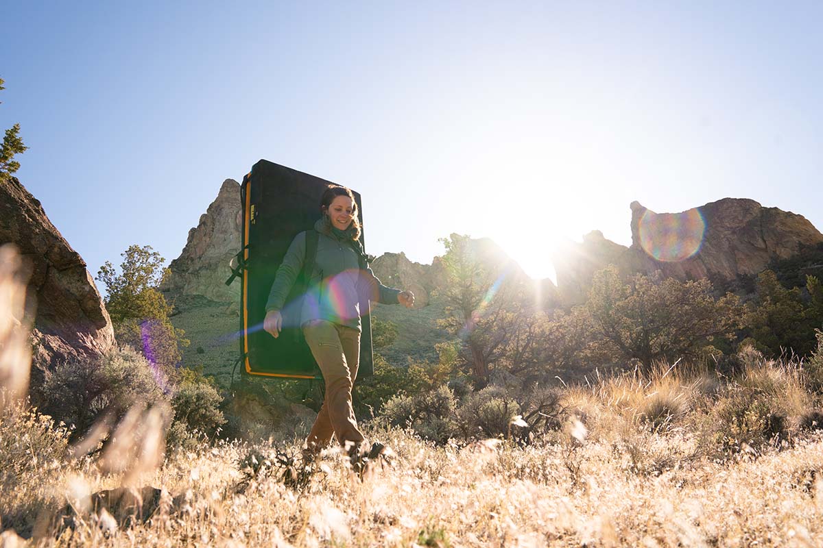 a woman hiking through a rocky landscape with a bouldering crash pad on her back