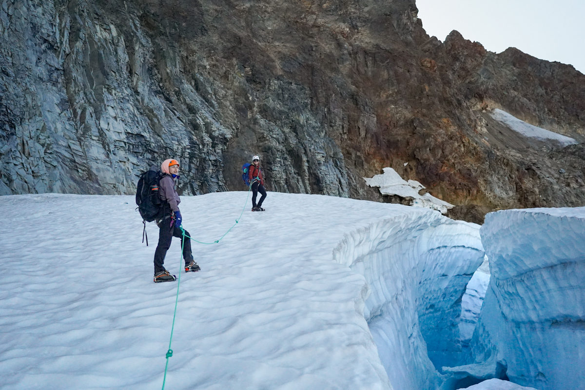 Two climbers use mountaineering boots and crampons to traverse a glacier field.