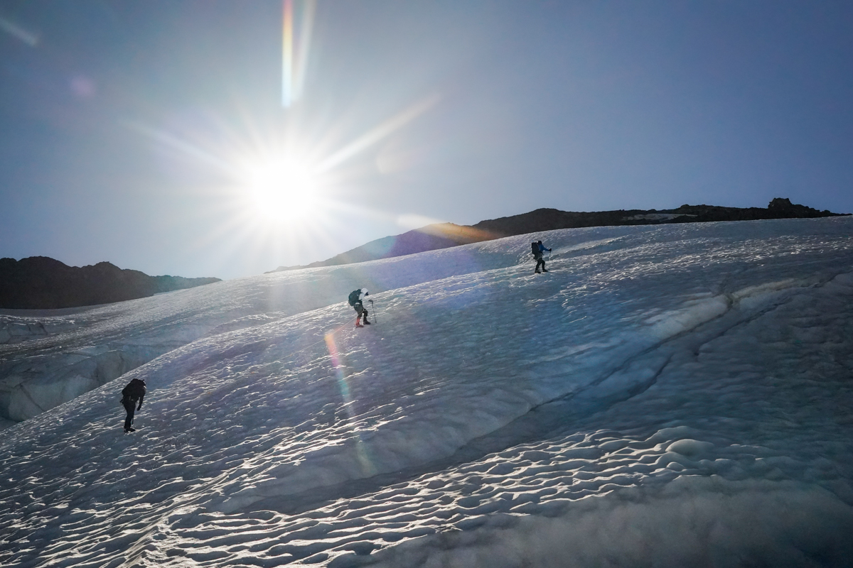 Three climbers traverse a glacier under the bright morning sun.