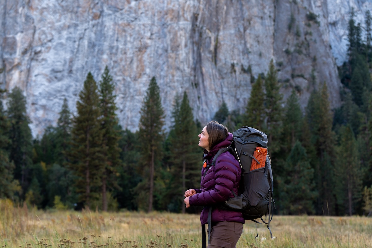 A woman with a large backpack stands in a valley looking up at the granite in Yosemite