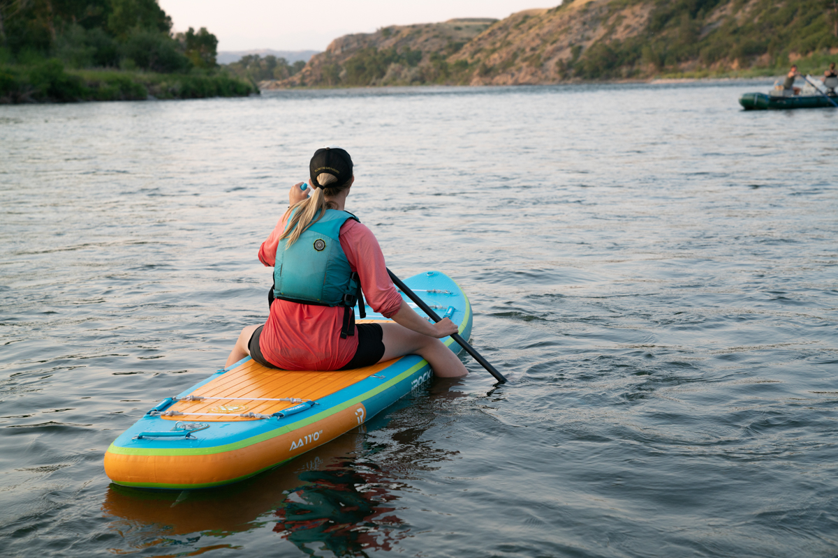 A woman sitting on a blue and orange paddle board on a river