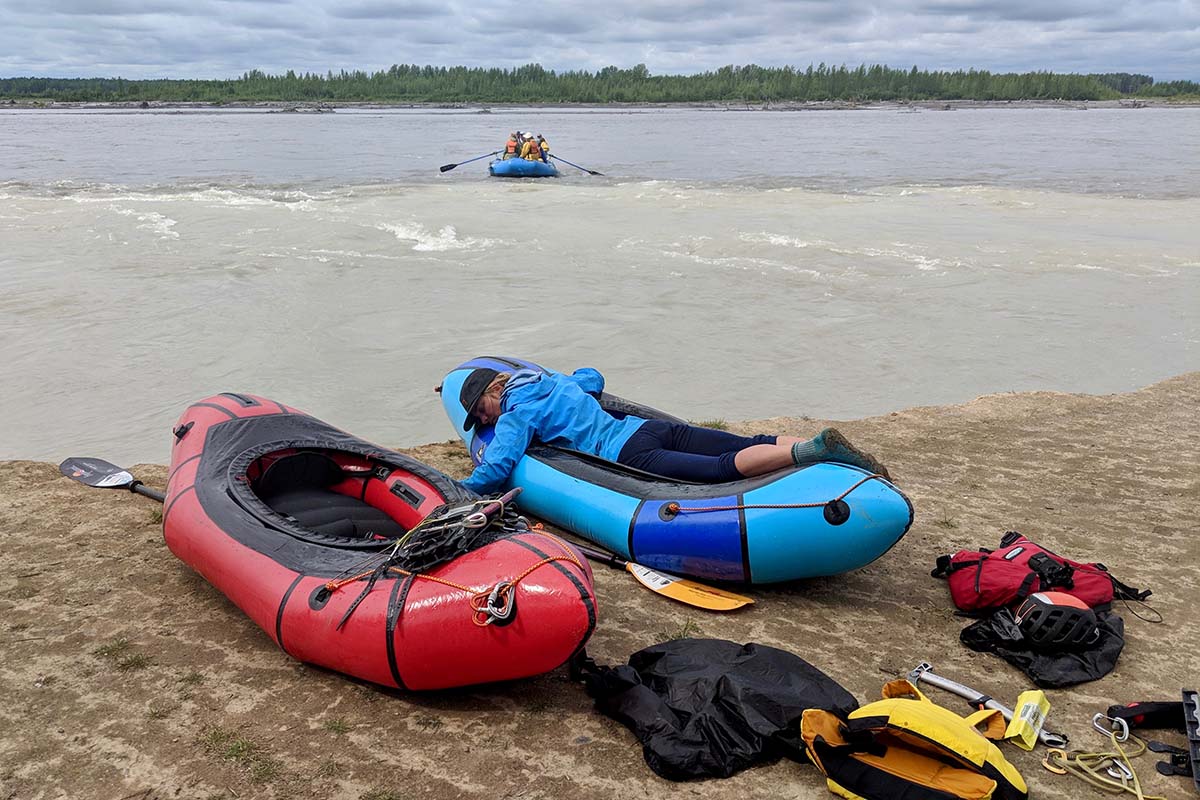 A woman laying in a blue packraft on the beach beside an empty red packraft 