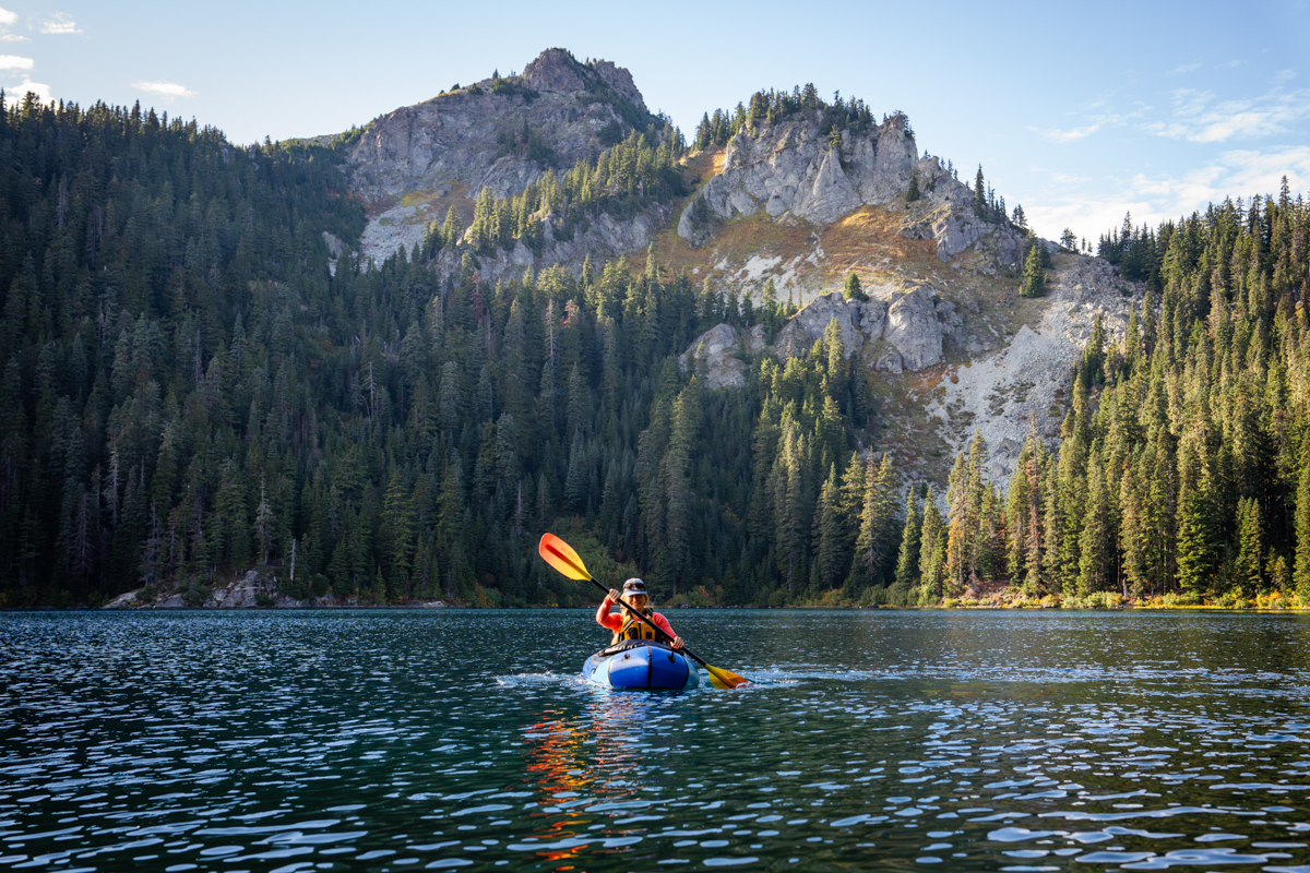 A woman paddling a blue packraft on a lake in front of a mountain