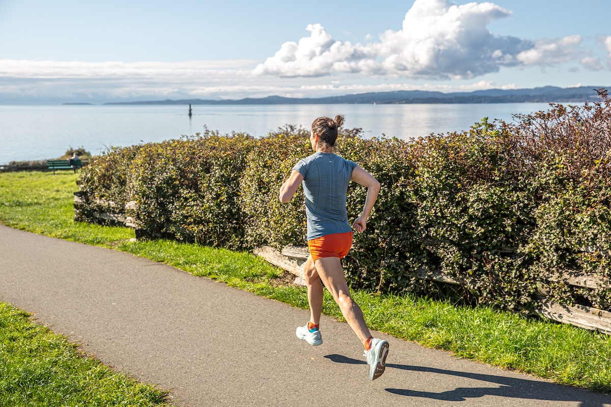 A woman is running from the camera as she tests shoes near the ocean on a paved pathway
