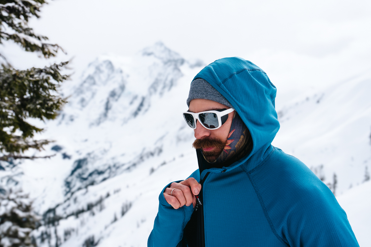 A man zipping up a blue baselayer with a snowy mountainous background
