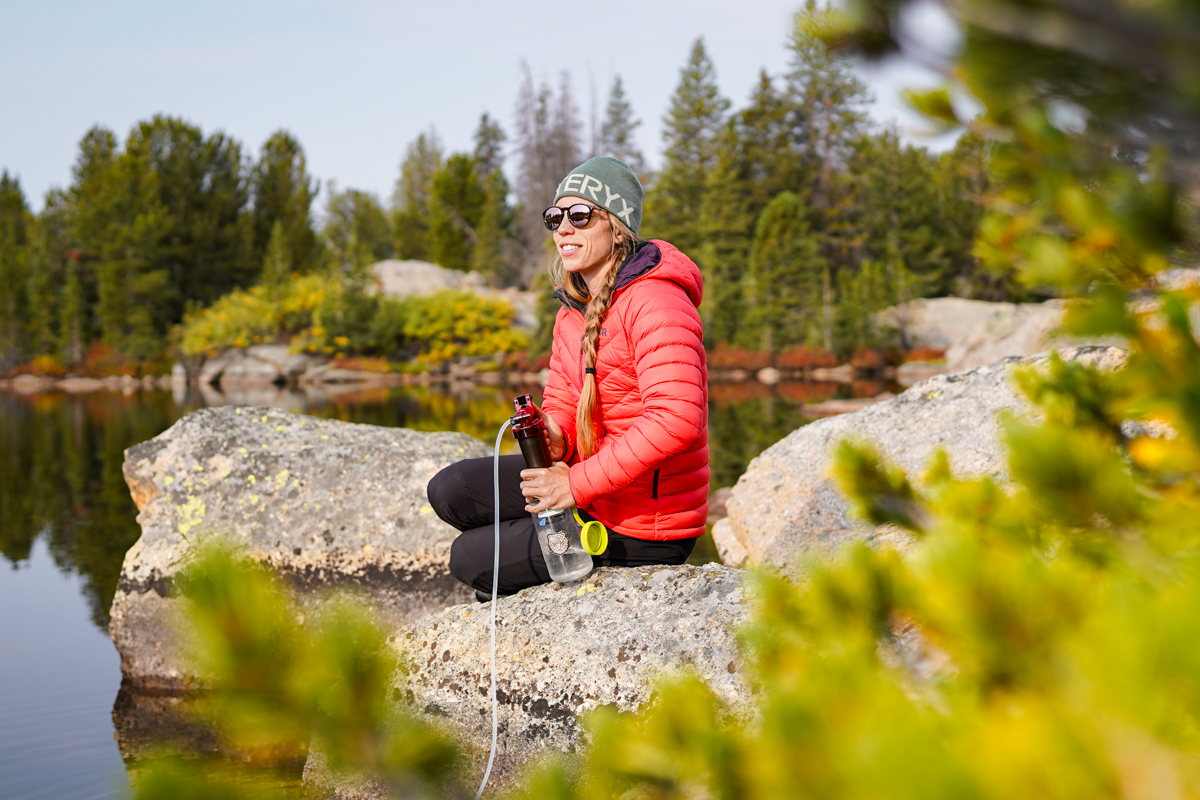 A woman in a brightly colored jacket sits on a rock and filters water into a Nalgene bottle