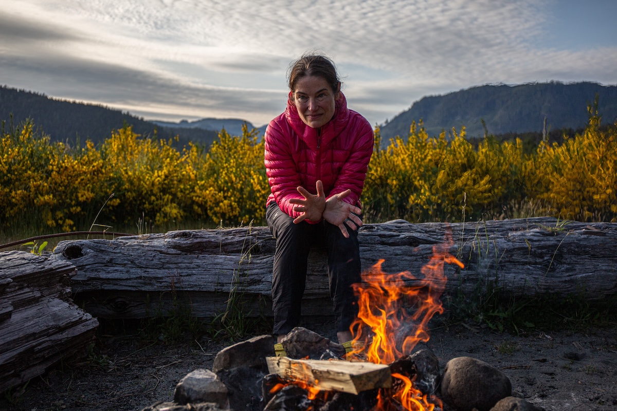 A woman sits by a fire wearing the Rab Mythic Alpine Light jacket