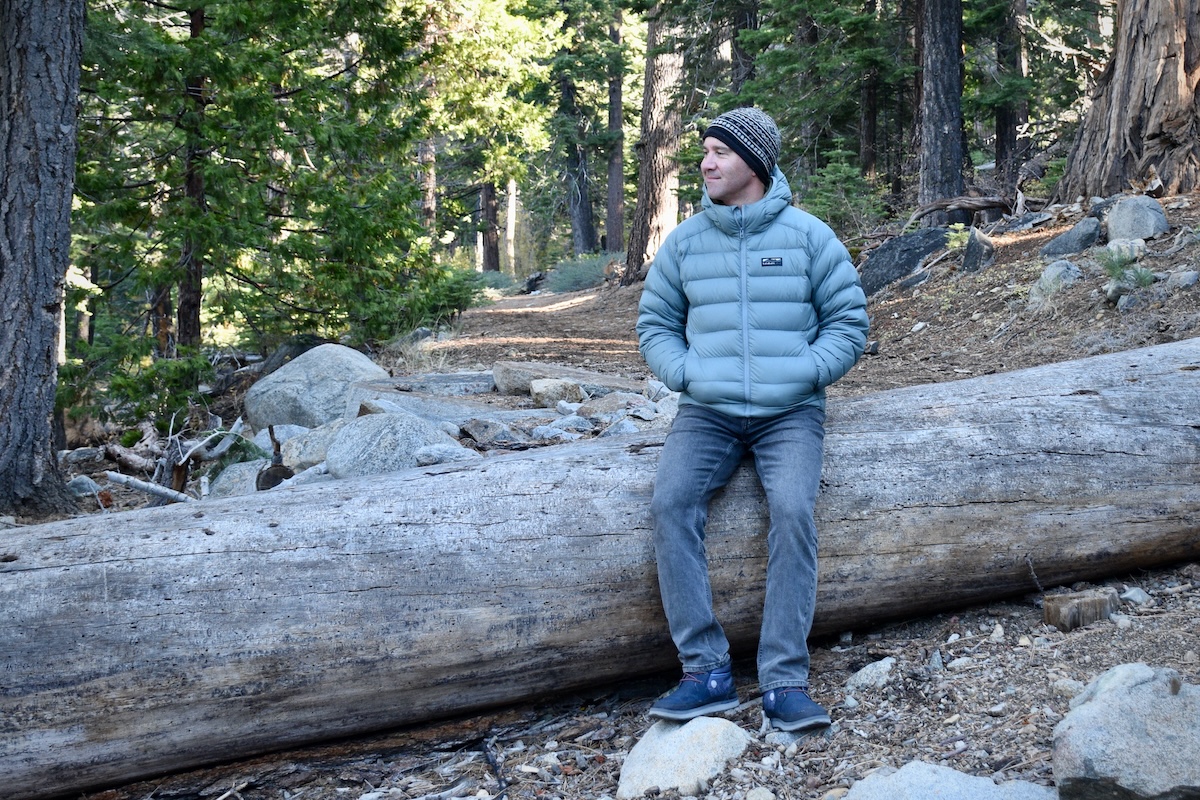 A man sits on a tree wearing the Tincup down jacket