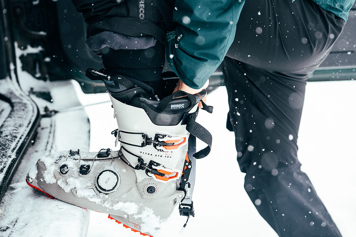 A skiier leans on the bumper of their car to get their ski boot on. 