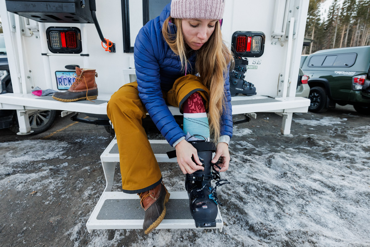 A woman sits next to her truck and puts on her ski boots.