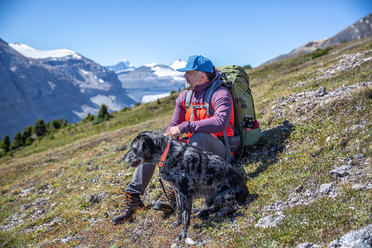 A man with a backpack sits on a grassy hill with his dog, admiring a mountain range