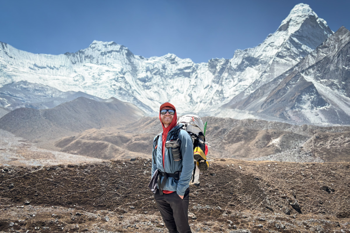 A man with a backpack smiles as he stands in front of an expansive snowy mountain range