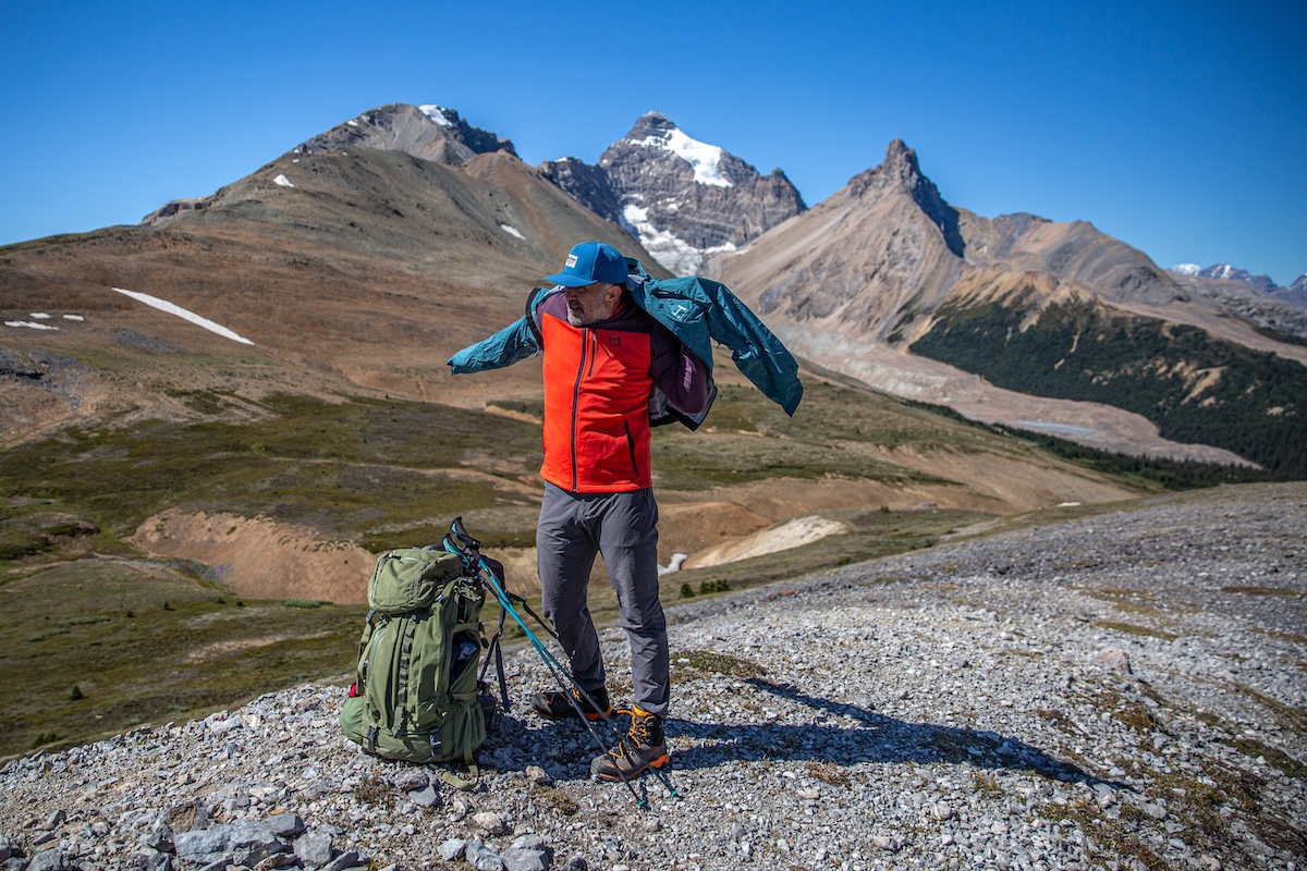 A man puts on a hardshell jacket with mountains in the background