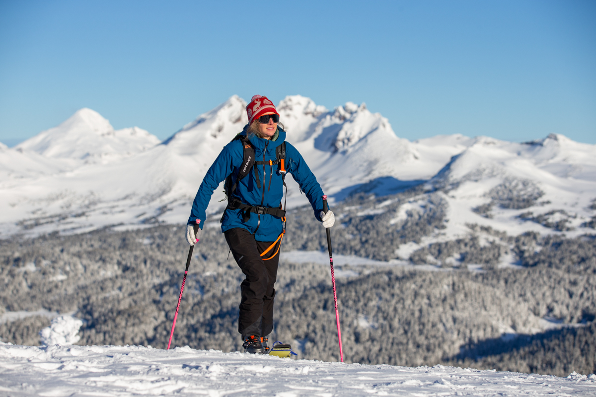 A woman skinning up a snowy slope wearing black ski bibs