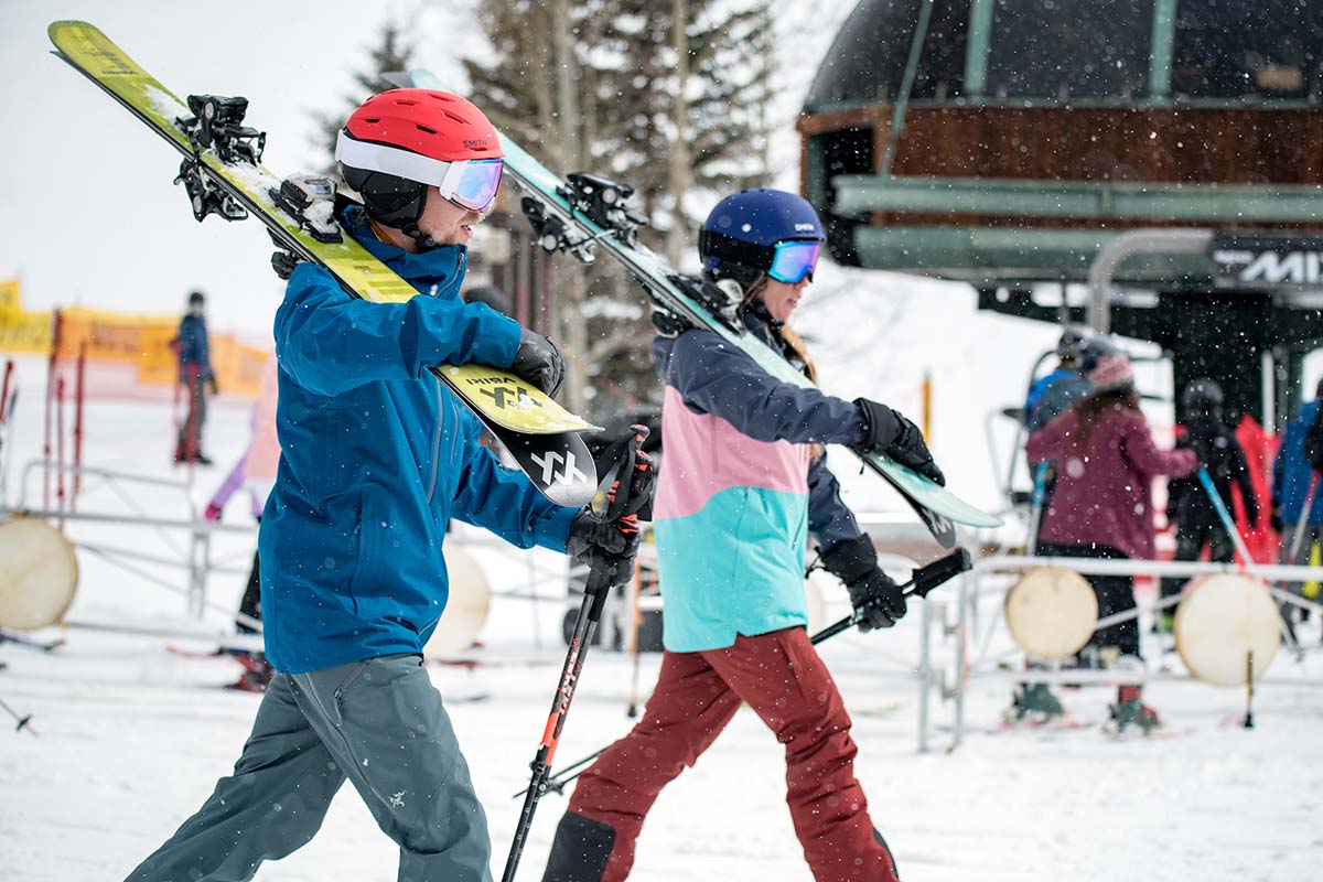 Two skiers walk by a ski lift at a ski resort carrying their skis on their shoudlers
