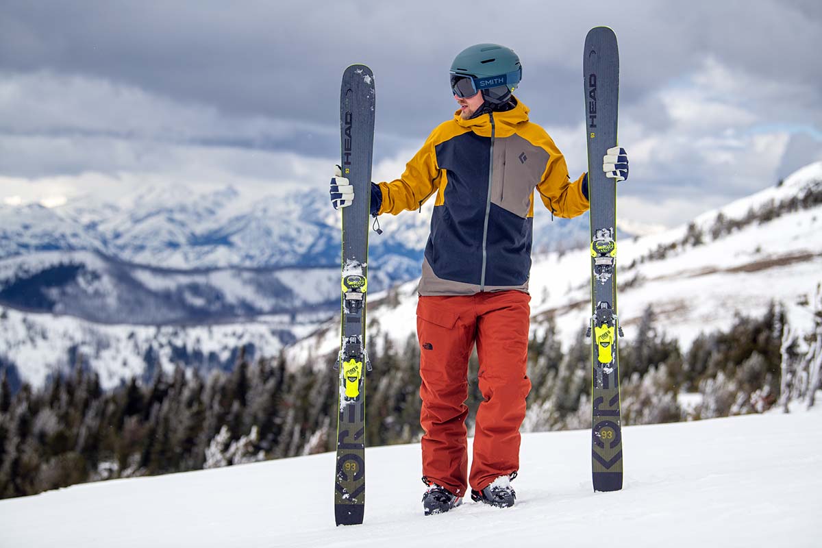 A skier stands at the top of a hill holding one ski in each hand to show the bindings