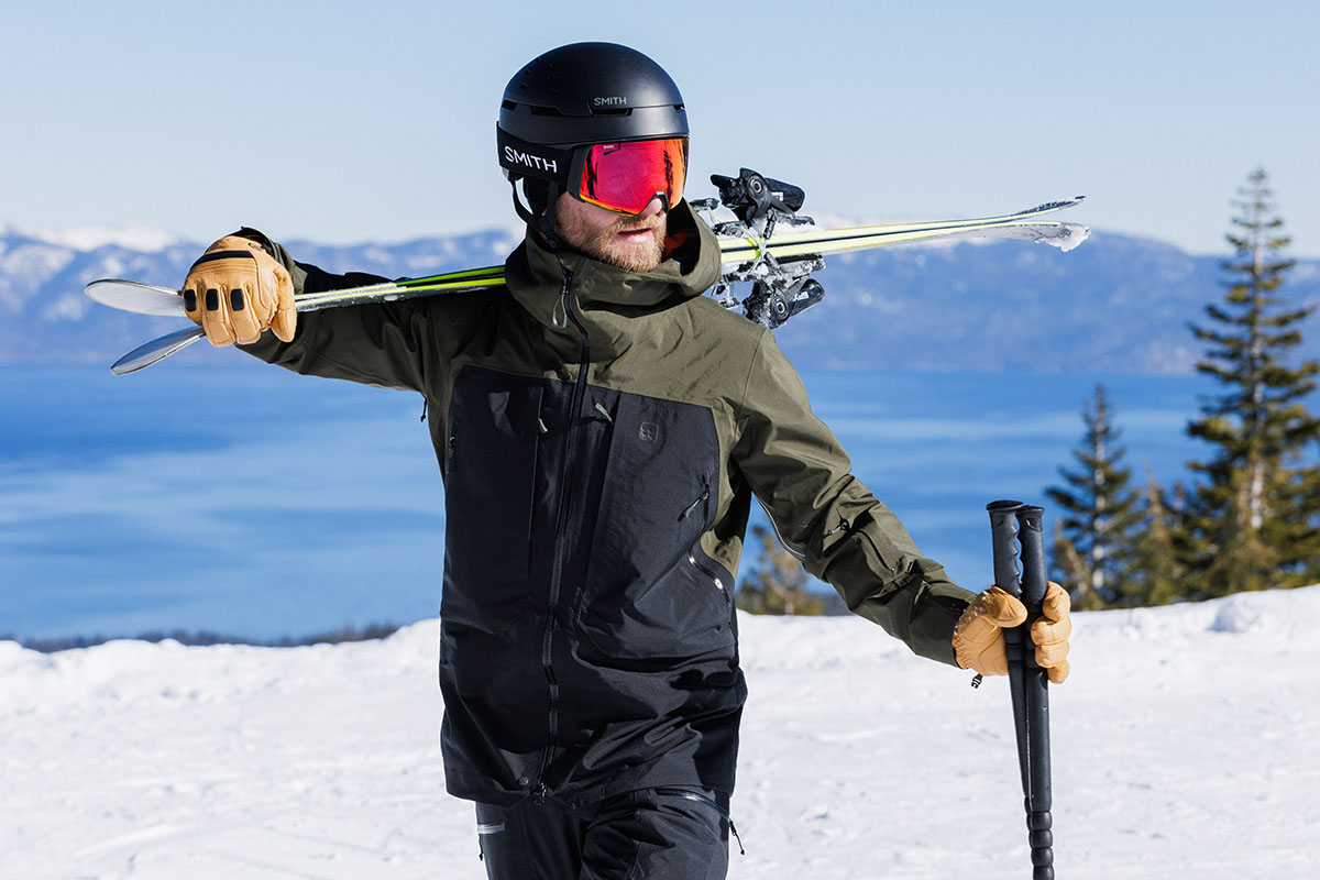 A man wearing a Smith helmet and goggles carries skis on his shoulder